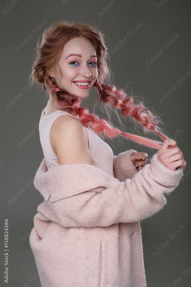 Portrait of a girl with pink hair in braids with blue and pink makeup, a pink coat, worn off the shoulder, standing in a half turn and pulling the tips of your braids, looking at the camera, smiling