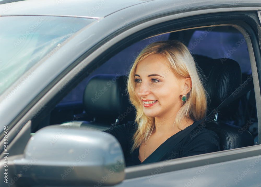beautiful blonde smiling behind the wheel of a grey car.