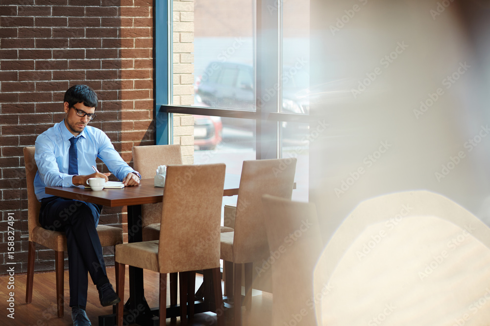 Concentrated bearded entrepreneur taking necessary notes while sitting in cozy cafe with panoramic windows, full length portrait Concentrated bearded entrepreneur taking necessary notes while sitting in cozy cafe with panoramic windows, full length portrait