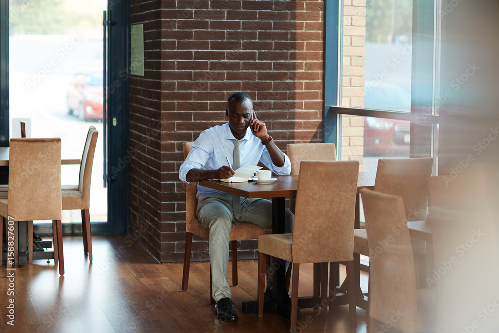 Full length portrait of concentrated African American entrepreneur talking to his business partner on mobile phone and taking necessary notes while sitting in lovely cafe Full length portrait of concentrated African American entrepreneur talking to his business partner on mobile phone and taking necessary notes while sitting in lovely cafe