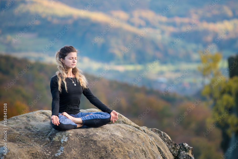Happy slim girl is practicing yoga and doing asana Siddhasana on the top of the mountain. Autumn forests, rocks and hills on the background