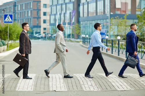Multi-ethnic group of confident coworkers crossing road in city center, modern office buildings on background, profile view