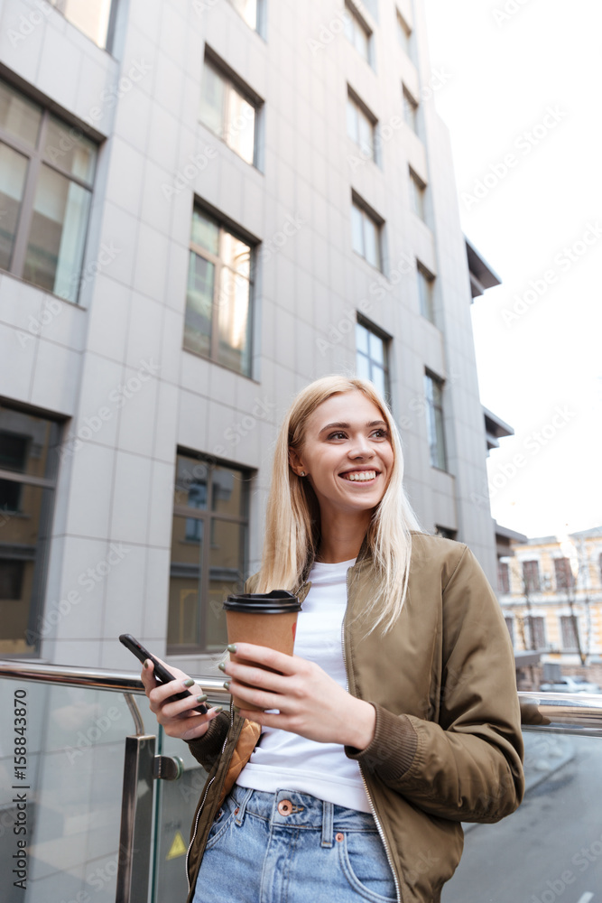 Fototapeta premium Cheerful woman with cup of coffee and smartphone looking aside