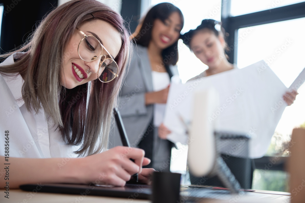 young designer drawing on graphics tablet while her colleagues looking at blueprint young designer drawing on graphics tablet while her colleagues looking at blueprint