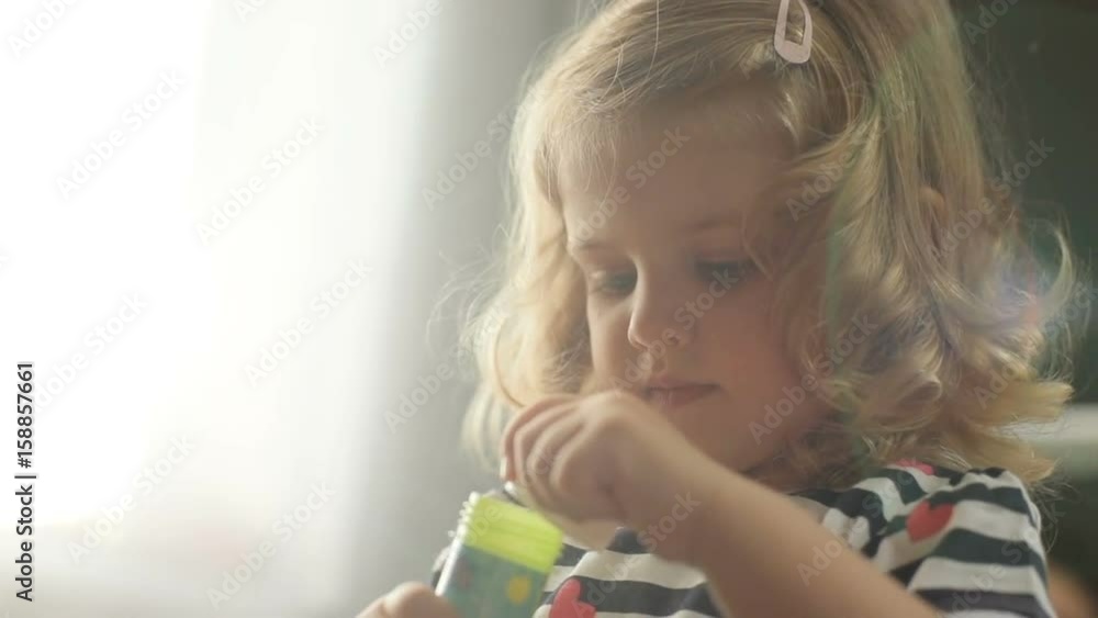 Portrait of side view of pretty caucasian young girl with blond curly hair making bubble blower on living background. Indoor. Portrait of side view of pretty caucasian young girl with blond curly hair making bubble blower on living background. Indoor.