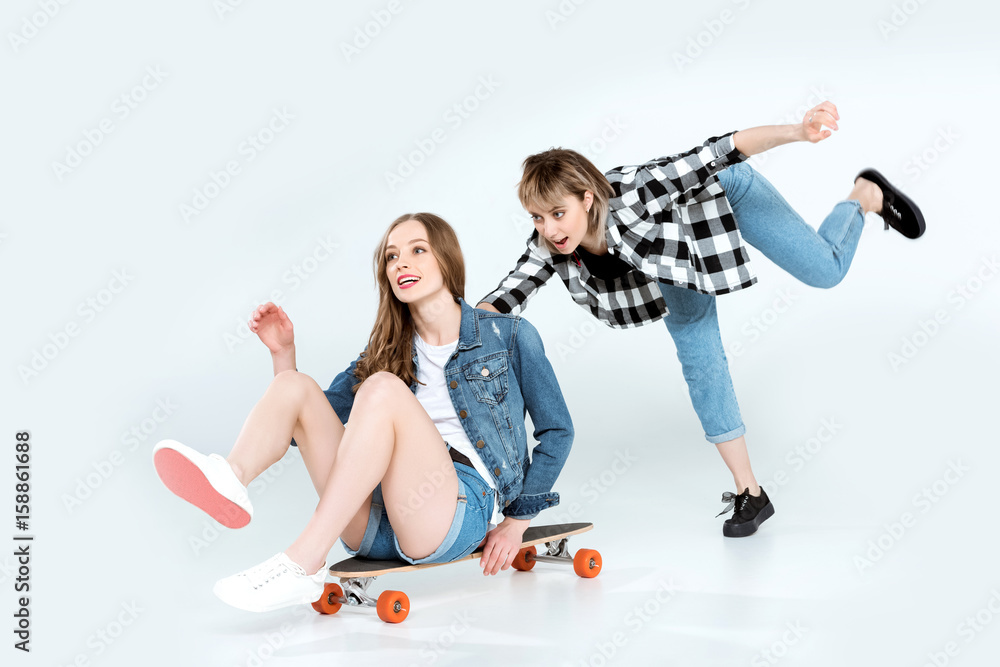 Attractive happy young girlfriends having fun with skateboard isolated on grey