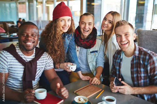 Group portrait of smiling friends looking at camera while drinking delicious coffee and chatting with each other in cozy small cafe