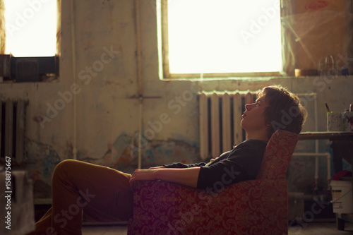 Young man sitting on vintage armchair in artist studio gazing upward photo