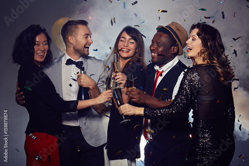 Happy friends clinking champagne flutes together while celebrating Christmas in nigh club, colorful confetti falling
