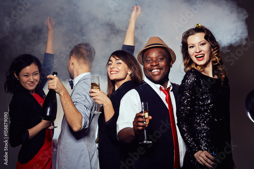 Enthusiastic young friends in stylish clothes dancing and drinking champagne in night club, thick smoke on background