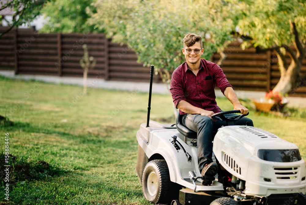 Fototapeta premium Smiling man riding a lawnmower and doing landscaping works