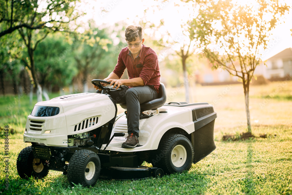 Fototapeta premium Ride-on lawnmower tractor with worker doing landscaping works at weekend sunset