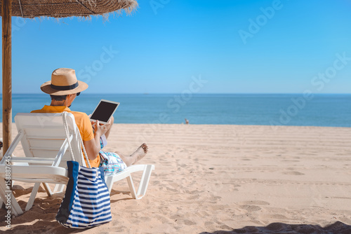Back view of tourist using tablet pc on the beach. Holiday relaxation vacation photography on sunny blue sky seacoast, sea shore outdoors background