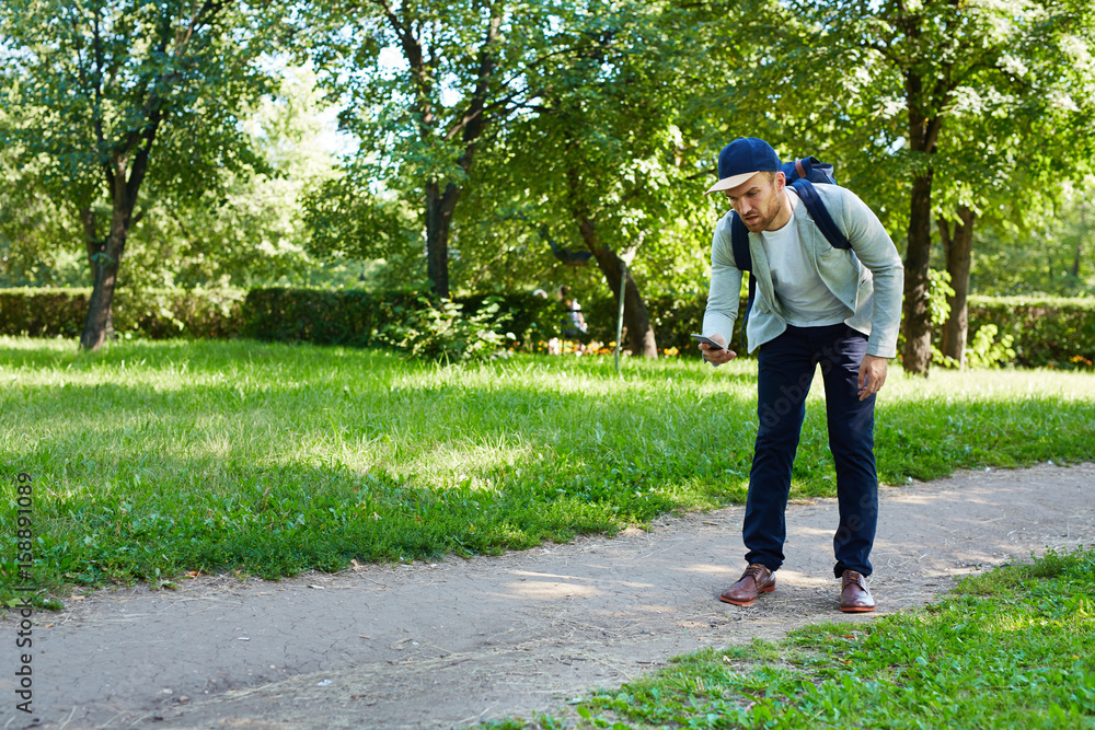 Fototapeta premium Concentrated young man walking down path and playing popular mobile game, full length portrait