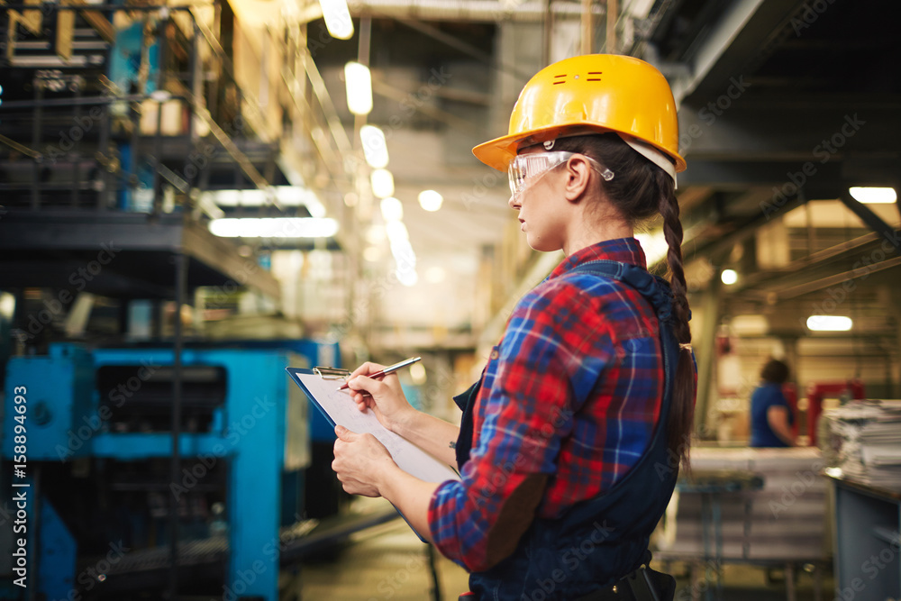 Profile view of beautiful young technician wearing protective goggles and helmet examining equipment in manufacturing plant Profile view of beautiful young technician wearing protective goggles and helmet examining equipment in manufacturing plant