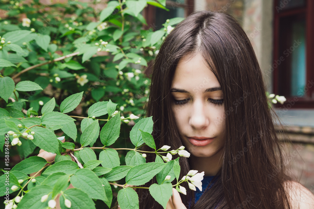 A very beautiful and cheerful girl stands on the street near a bush with white flowers A very beautiful and cheerful girl stands on the street near a bush with white flowers