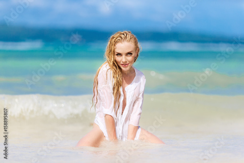 Happy beautiful blonde woman with long hair dressed in white on a tropical beach. Blue sea in the background. Summer vacation concept.
