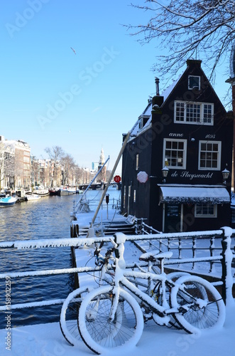 Typical two Dutch bikes covered by snow surrounded by river and historical Dutch house. Sunny winter romantic day. February in Amsterdam.