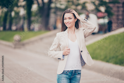 Young attractive model is posing walking outdoors at the spring street in a formal beige jacket, jeans and white t shirt, fixing her hair, smiling © deagreez