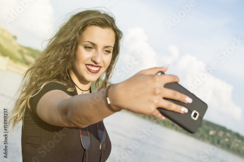 Beautiful girl taking a selfie. Beautiful day by the river. Lens flare in the background.