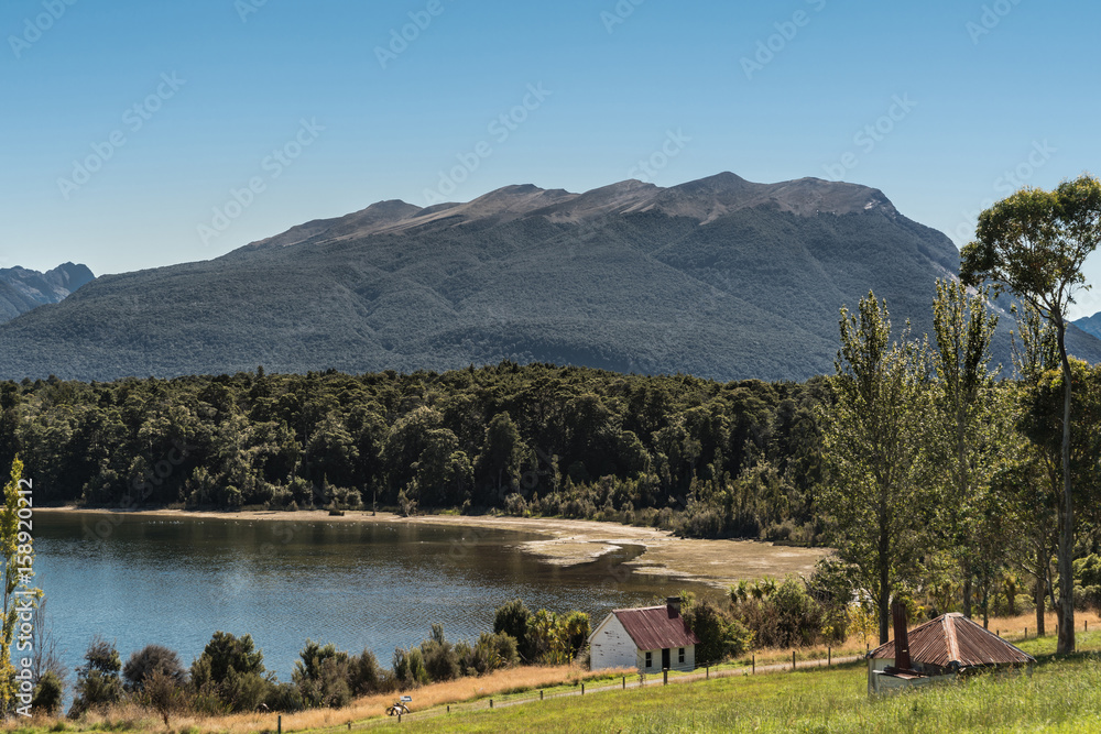 Te Anau, New Zealand - March 16, 2017: shoreline of the lake shows forests and small farm building, now a cottage. Green and yellow grass in foreground, dark mountains in back under blue sky.