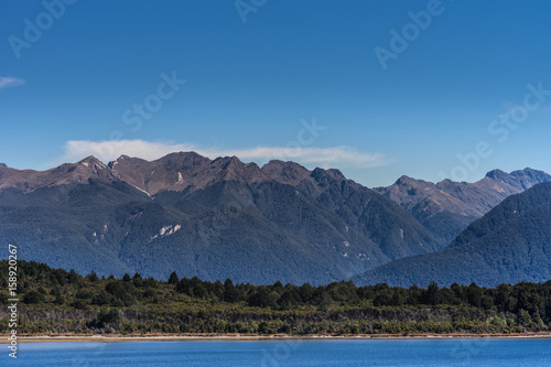 Te Anau, New Zealand - March 16, 2017: Looking over part of majestic lake Te Anau with dark blue water and sky. Big mountainous slopes with forests and other green vegetation. Minimal white clouds.