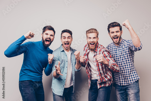 Yes! Winners! Four cheerful young men are standing and gesturing for the victory on pure background in casual outfit and jeans. They are fans of sports games as football, basketball, hockey, baseball photo