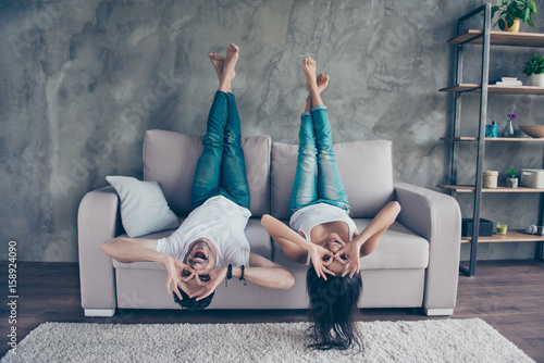 Funny couple with glasses gesturing is lying upside-down on the sofa at home. They are so cheerful, having fun together, go crazy, brother and sister photo