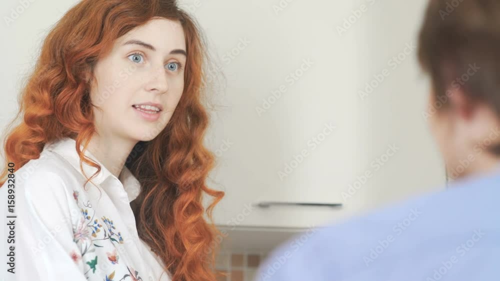 Man and a girl drink coffee together and talk in the kitchen in a bright interior. A young woman with blue eyes and bright red hair communicates with her man. Home interior. Dialogue at breakfast. Man and a girl drink coffee together and talk in the kitchen in a bright interior. A young woman with blue eyes and bright red hair communicates with her man. Home interior. Dialogue at breakfast.
