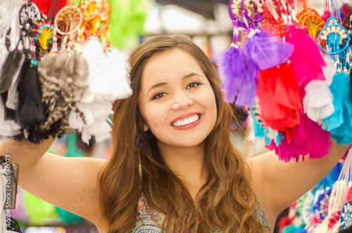 Close up of a beautiful smiling young woman hiding behind a colorful catchdreamer, in colorful market fabrics background