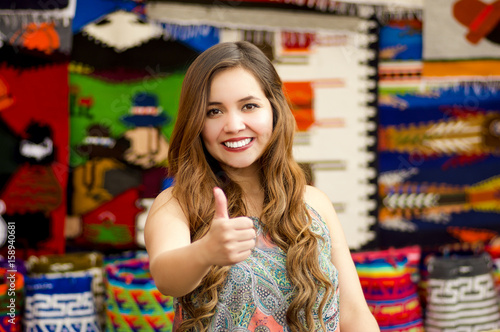 Beautiful smiling young woman with humbs up and posing for camera, with colorful andean traditional clothing fabrics background