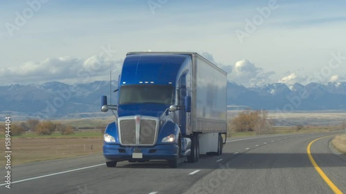 CLOSE UP: Freight semi truck transporting goods driving along the multiple lane highway past pasture fields and farmlands. Lorry trucking load on sunny day with Rocky Mountain Range in the backgrounds