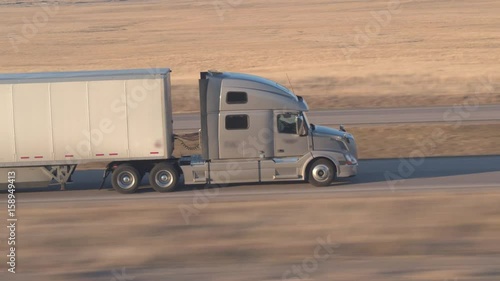 AERIAL, CLOSE UP Freight container semi truck hauling goods driving along the empty interstate highway through the prairie fields in the Great Plains, America. Trailer shipping cargo on sunny evening