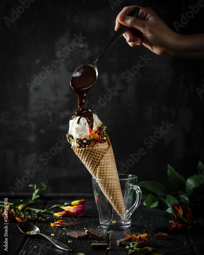 ice cream cone in a glass cup with woman's hand holding a spoon of melted chocolate pouring on ice cream on a black background, low light