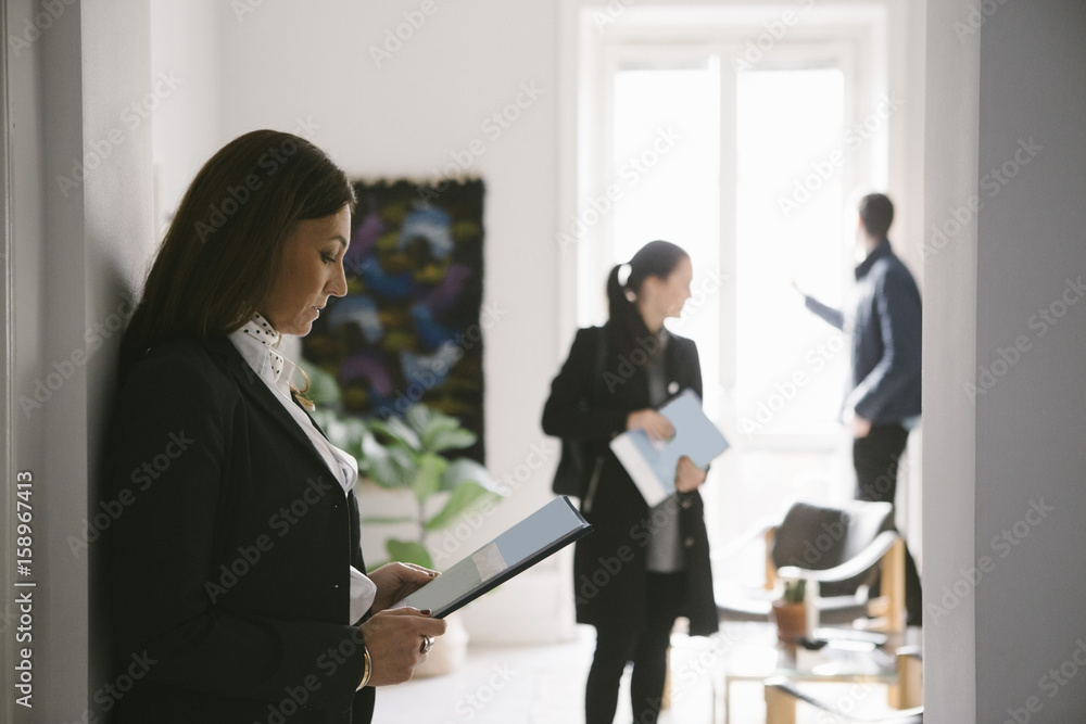 Side view of female realtor reading brochure with couple in background Side view of female realtor reading brochure with couple in background
