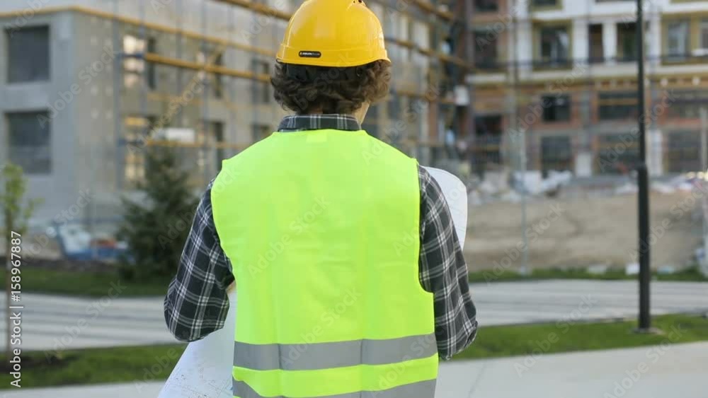 Back view of caucasian architect in yellow helmet and green vest looking drawing standing on building background. Outdoor. Back view of caucasian architect in yellow helmet and green vest looking drawing standing on building background. Outdoor.