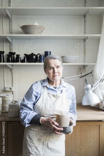 Portrait of senior female potter standing with pitcher against shelves at workshop photo