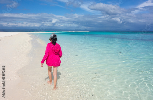 Back of young Asian girl in pink jacket walking on perfect white sand beach and clear blue sea. Summer outdoor nature holiday serenity. Kalanggaman Island, Philippines. Background, copy space.