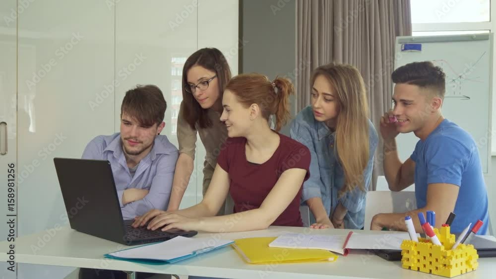 Five creative young people showing teamwork at the office. Handsome caucasian guys sitting at the table from the both sides of three girls. Two pretty women giving some advices to their female Five creative young people showing teamwork at the office. Handsome caucasian guys sitting at the table from the both sides of three girls. Two pretty women giving some advices to their female