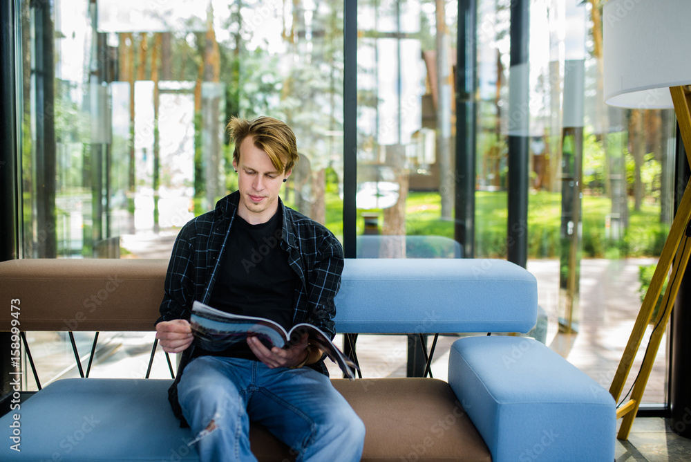 Handsome young man with blond hair sitting and reading a magazine Handsome young man with blond hair sitting and reading a magazine