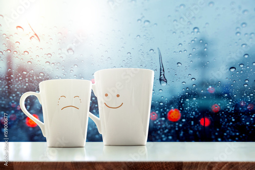 Encouragement concept, Friend of Coffee Mug with Sadness crying face cartoon and kindness happy face inside the room, Blurred city lights and rain drop in city as outside view through glass window photo