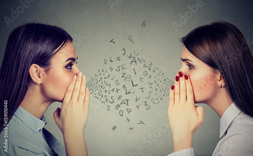 Two women whispering a gossip secret to each other with alphabet letters in-between