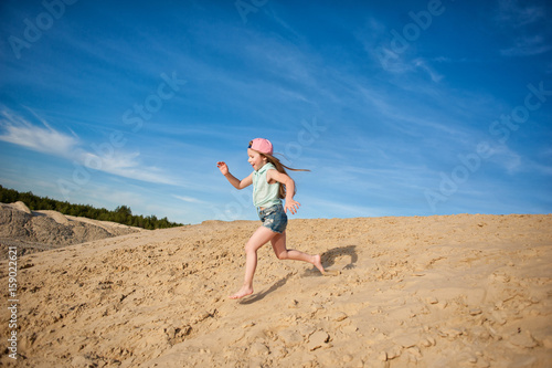 Little active girl walking, running, jumping, having fun along the sand on the beach against the background blue sky in summer vacation