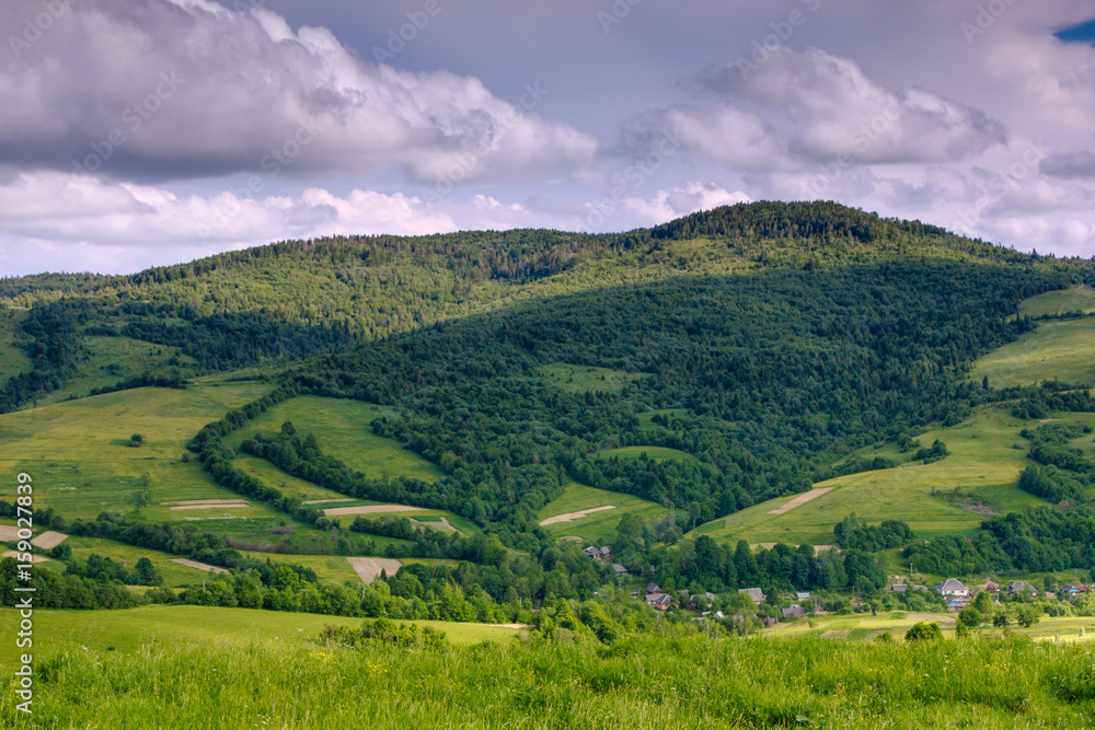 Obraz premium Scenic landscape of the countryside near alpine mountains. View of green hills covered by trees. Summer natural background. Sunlight shines through the trees.