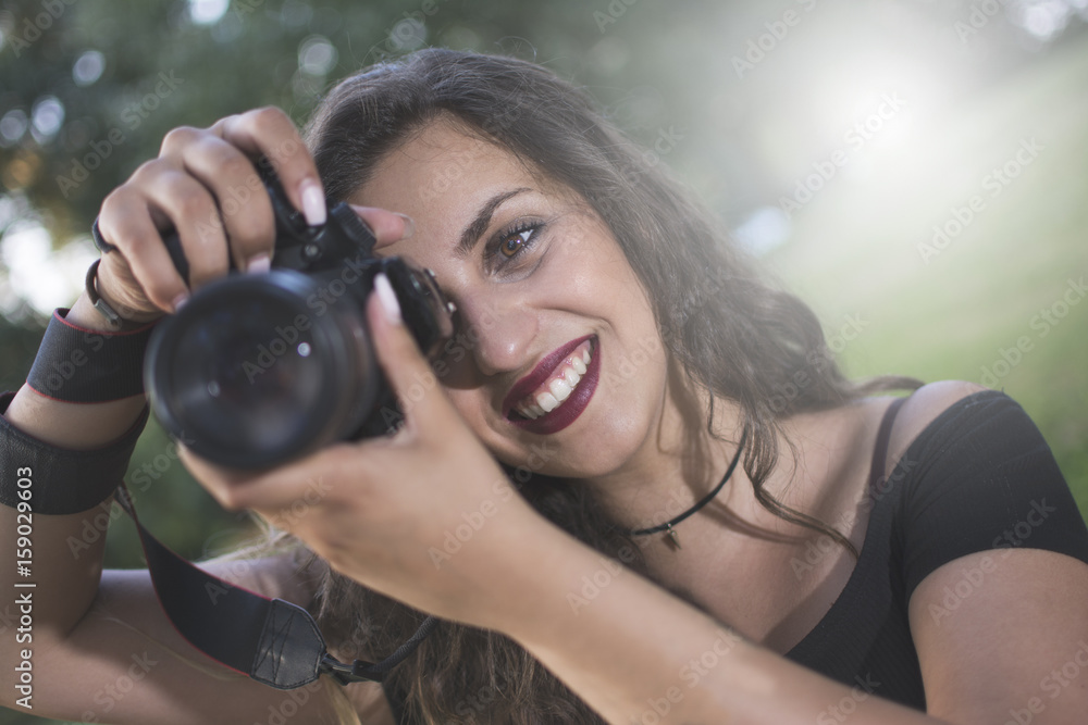 Fototapeta premium Woman photographer with a professional camera taking a photo. Beautiful day by the river. Lens flare in the background.