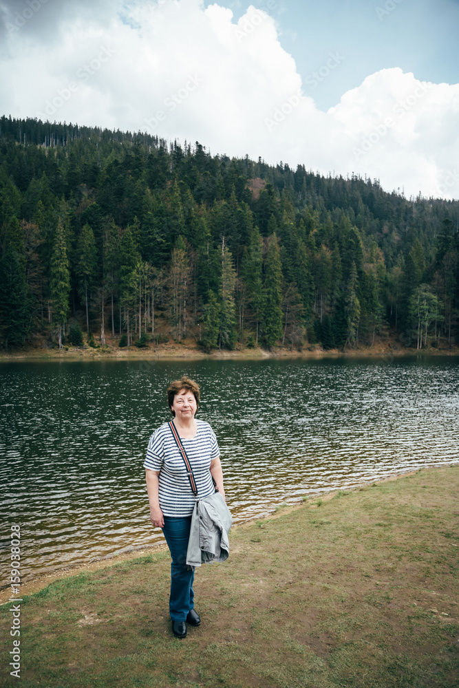 Senior beautiful woman standing on the bank of the mountain lake surrounded by forest. Travel concept