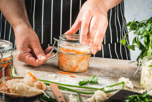 Concept of fermented meal. Home canned food and billets. Vegan food. Vegetables. Person (woman) cooking sauerkraut. Cans with conservation, broccoli, carrots, greens, onions. photo