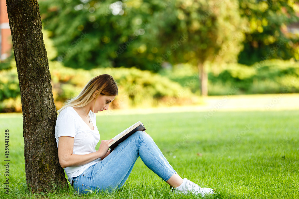 Modern student sitting in park under the tree and reading the book