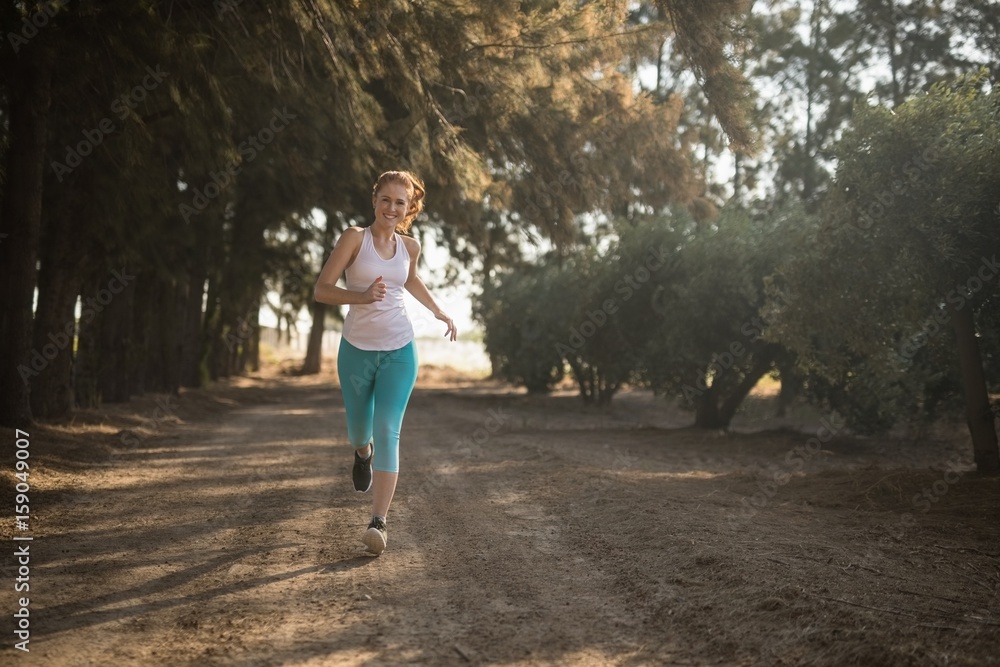 Smiling young woman jogging on dirt road during sunny day