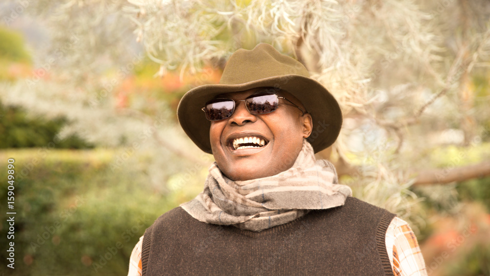 Handsome and happy man enjoying the outdoors. Portrait of smiling African-American man outdoors wearing hat and sunglasses hiking, traveling, or modeling. Trees in background unfocused, copy space.